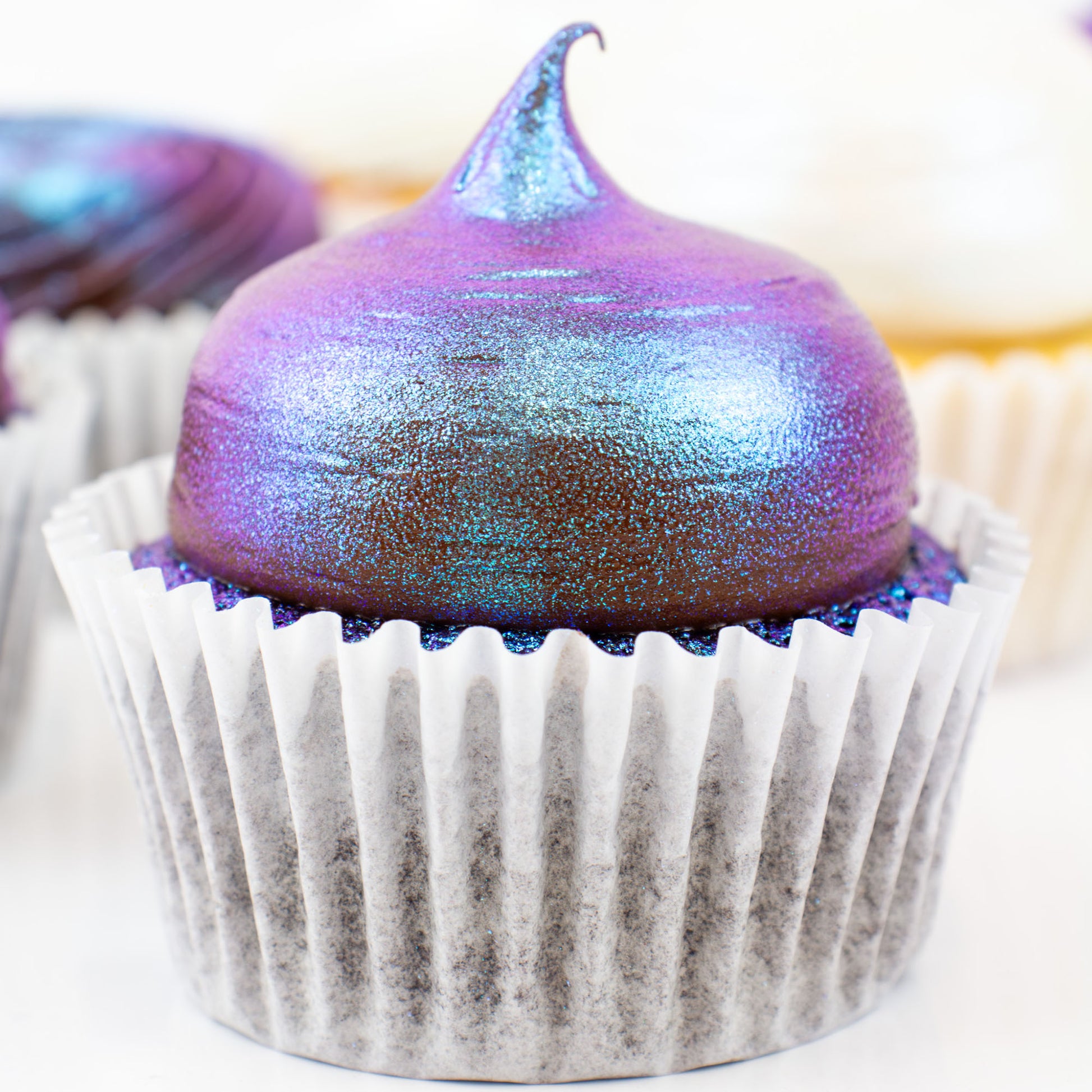 Cupcake with iridescent purple and blue frosting in a white paper cup.