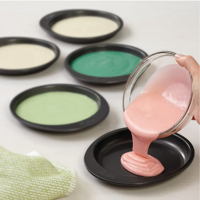 Pink cake batter being poured into a cake pan with other coloured batters in molds on a white background.