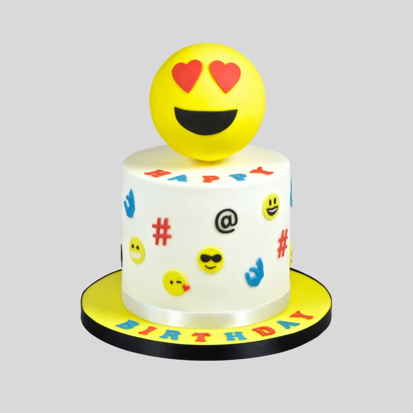 A custom birthday cake decorated with white icing and colorful emoji symbols, topped with a large yellow smiley face with red heart eyes, on a yellow and black round base with 'BIRTHDAY' written in colorful letters.