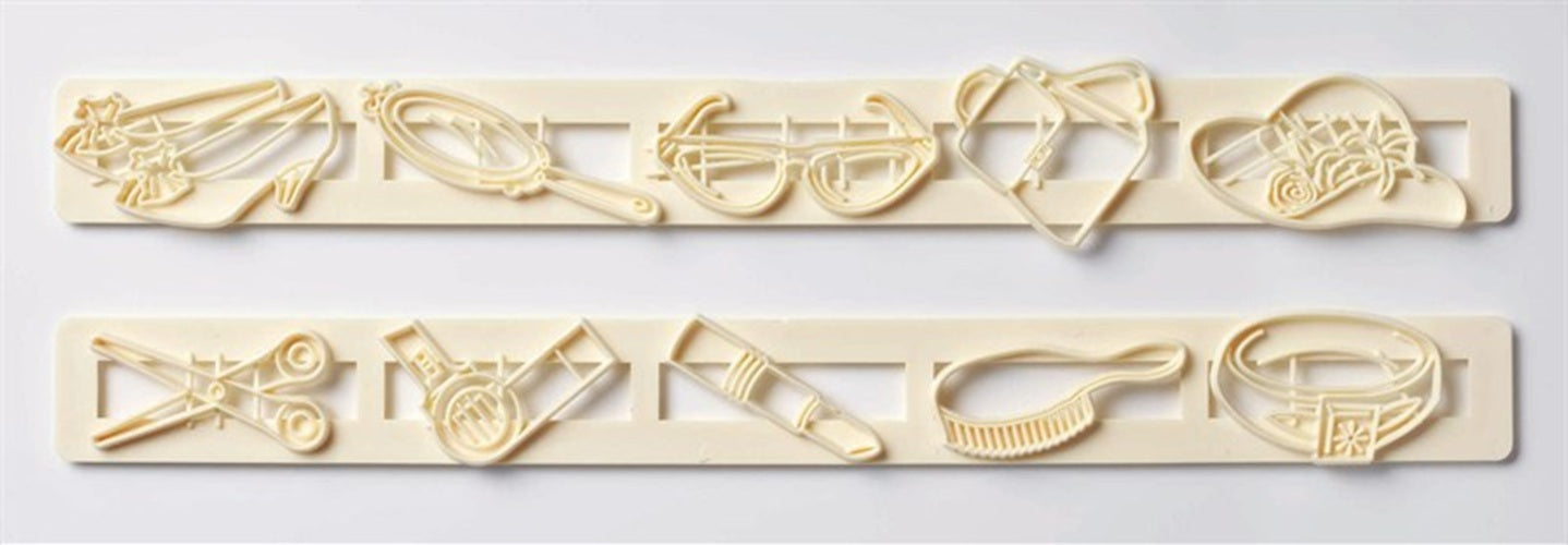 A set of cream-colored gumpaste icing cutters arranged in two rows on a light gray background. Including a ring, hair brush, lip stick, hair dryer, scissors, shoes, hand mirror, sunglasses, hand bag, and summer hat.
