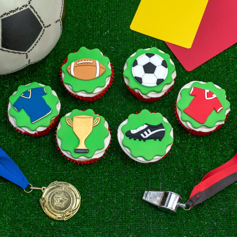 Cupcakes decorated with sports-themed designs on a green surface with sports equipment.