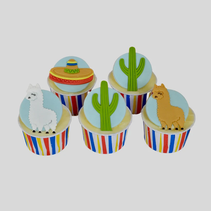 Four decorated cupcakes arranged on a light gray background. The cupcake liners have vertical red, blue, and yellow stripes, and the tops are decorated with light blue fondant, green cacti, white and tan alpacas, and a yellow sombrero.