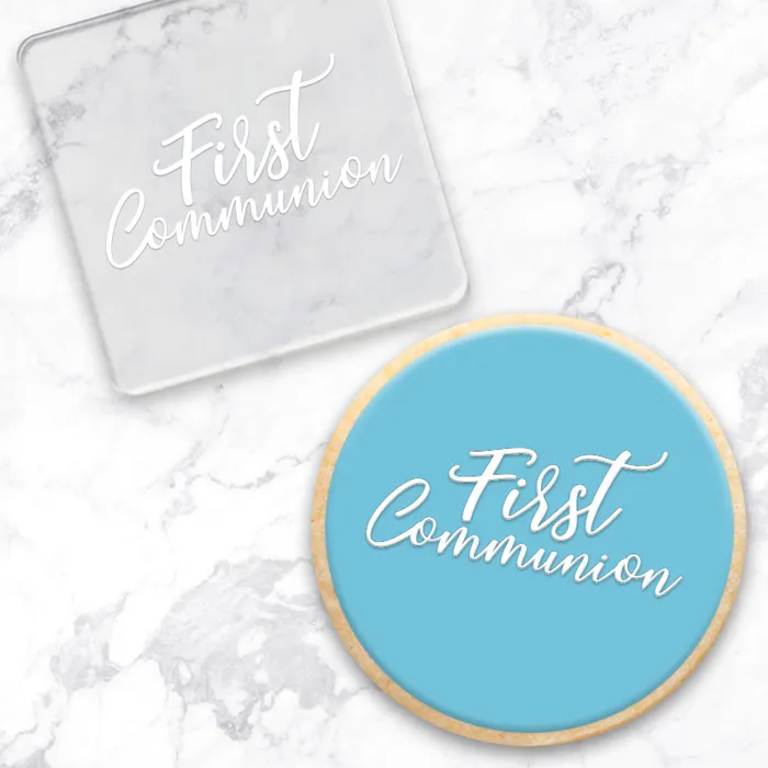 A square clear acrylic debosser and a round light blue cookie, both printed with the words 'First Communion' in white script, arranged on a white marble surface.