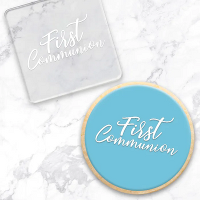 A square clear acrylic debosser and a round light blue cookie, both printed with the words 'First Communion' in white script, arranged on a white marble surface.