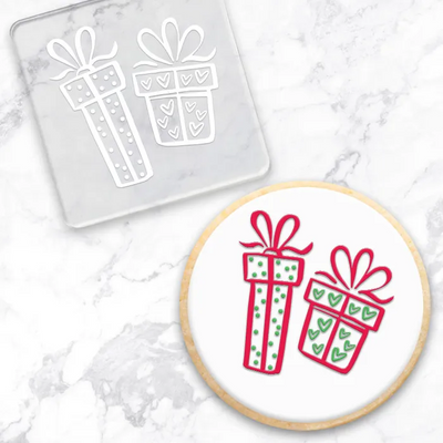 A round white cookie with two gift box illustrations in red and green icing, shown alongside a clear square acylic debosser with matching white gift box outlines on a light marble surface.