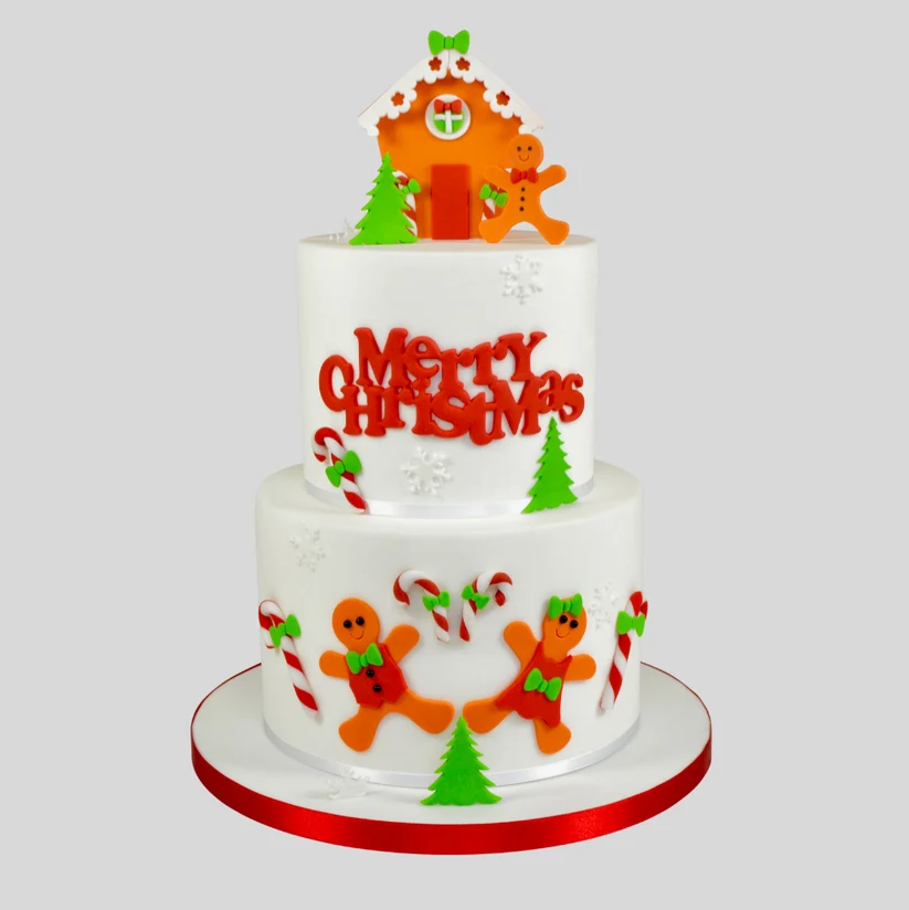 A two-tier white frosted Christmas cake decorated with orange gingerbread men, green trees, candy canes, and red 'Merry Christmas' lettering, topped with a gingerbread house and gingerbread man figure, on a light gray background.