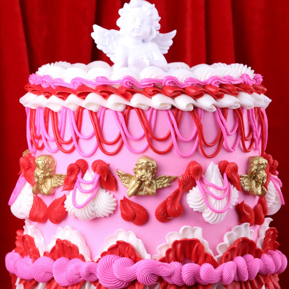 Decorative cake with pink and red icing, white and gold figurines against a red curtain background