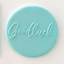 A round light blue decorative icing plaque featuring the word 'Goodluck' in raised cursive script, shown on a plain light background.