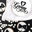 A styled flat-lay of groom-themed round decorations or cookies on a black surface with white confetti. The main piece is a white round with black script reading “Groom” and a tuxedo silhouette forming the letter “O.