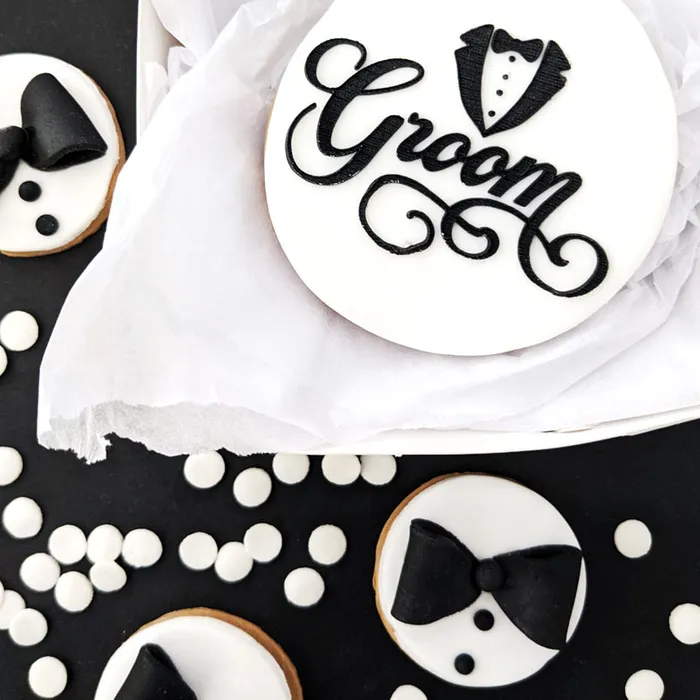A styled flat-lay of groom-themed round decorations or cookies on a black surface with white confetti. The main piece is a white round with black script reading “Groom” and a tuxedo silhouette forming the letter “O.