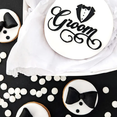 A styled flat-lay of groom-themed round decorations or cookies on a black surface with white confetti. The main piece is a white round with black script reading “Groom” and a tuxedo silhouette forming the letter “O.