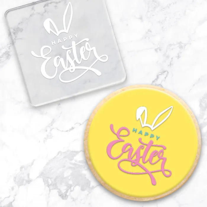 A clear acrylic debosser that reads Happy Easter, with a set of Bunny ears. Alongside a yellow finished cookie, made with the tool