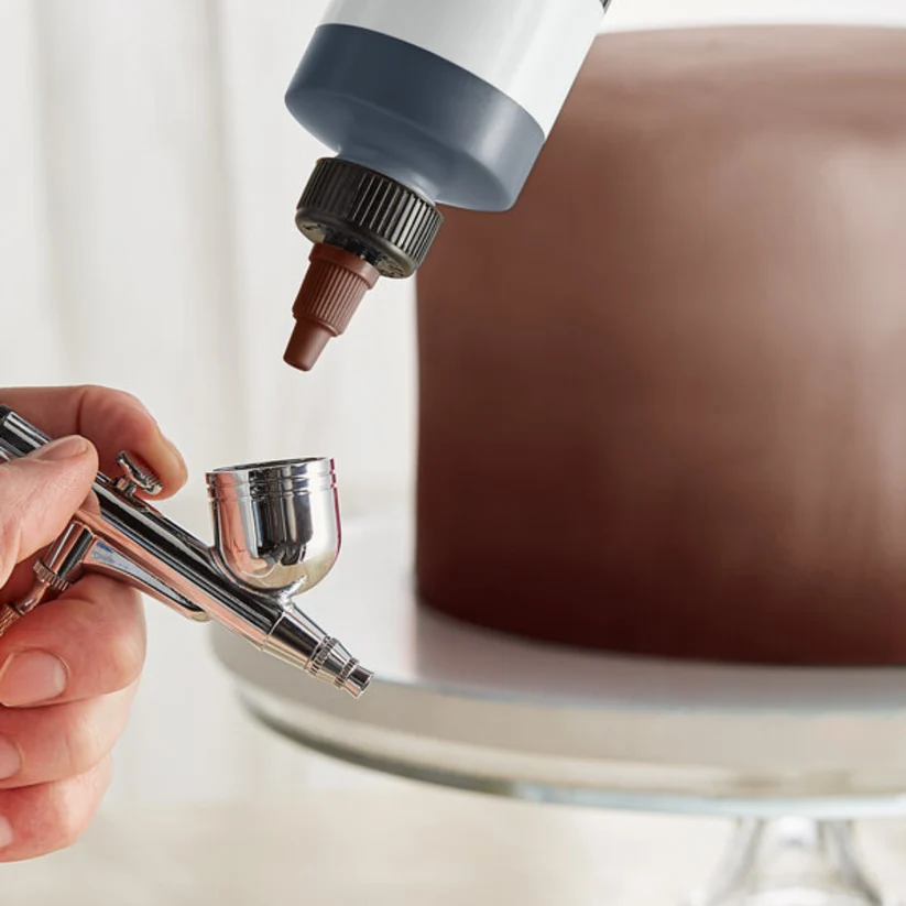 an airbrush with a brown cake in background on a cake pedestal