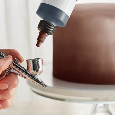 an airbrush with a brown cake in background on a cake pedestal