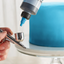 A close-up of a hand holding an airbrush tool while a bottle with a blue nozzle applies liquid to the cup of the airbrush. In the blurred background, a blue frosted cake sits on a cake stand.