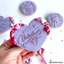 A hand holds a decorated heart-shaped Valentine's Day cookie in lavender purple icing with silver 'Happy Valentine's Day' script and pink, red, and white heart sprinkles. Other similar decorated cookies are arranged on a white background.