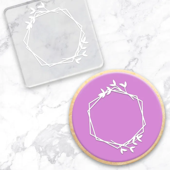 A white geometric wreath with leafy branches design shown on a round purple cookie and a clear square acrylic debosser, placed on a white marble surface.