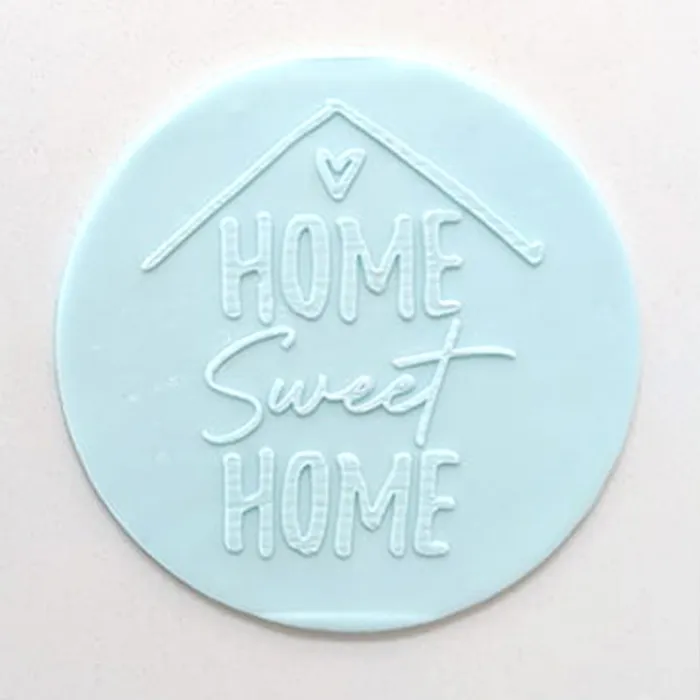 A round, light blue fondant plaque featuring the embossed text 'HOME Sweet HOME' with a small house outline and heart at the top, set against a plain off-white background.