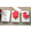 Three whitefoam pads on a neutral surface, showing red fondant icing duck shape being moulded into one mould and another mold with a partially formed duck. A white rolling pin and a small hand tool are visible, along with a person's fingers holding one mold on the left.