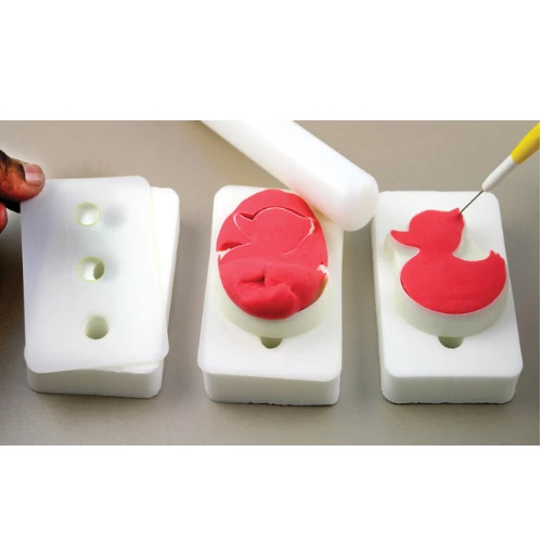 Three whitefoam pads on a neutral surface, showing red fondant icing duck shape being moulded into one mould and another mold with a partially formed duck. A white rolling pin and a small hand tool are visible, along with a person's fingers holding one mold on the left.