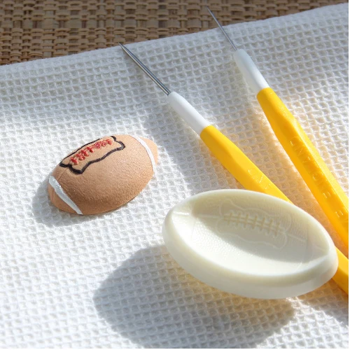A football-themed pop it mould cutter by Jem alongside a fondant example of a small brown rugby football with 'PARTY GO!' written on it, and two yellow-handled metal tools, all arranged on a white textured mat.