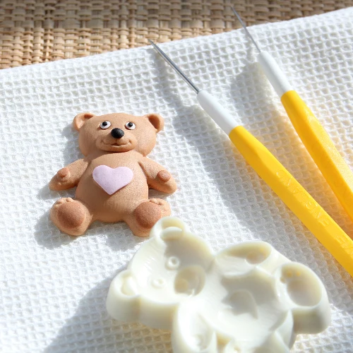 Small teddy bear fondant figurine with a heart on a textured white surface with yellow tools.