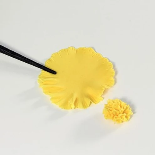 A yellow flower-shaped sugar paste piece is shown on a white background, with a black tool touching the edge of the flat piece and a smaller textured flower piece beside it.