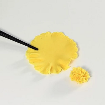 A yellow flower-shaped sugar paste piece is shown on a white background, with a black tool touching the edge of the flat piece and a smaller textured flower piece beside it.