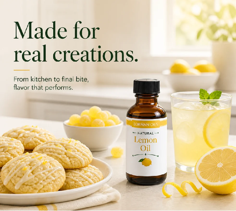 A bottle of Lorann's Natural Lemon Oil stands on a kitchen counter beside a glass of lemonade, cookies, lemons, and decorative text reading 'Made for real creations.'.