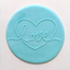 A round light blue fondant plaque featuring the word 'love' in a cursive script that forms a heart outline, set against a plain off-white background.