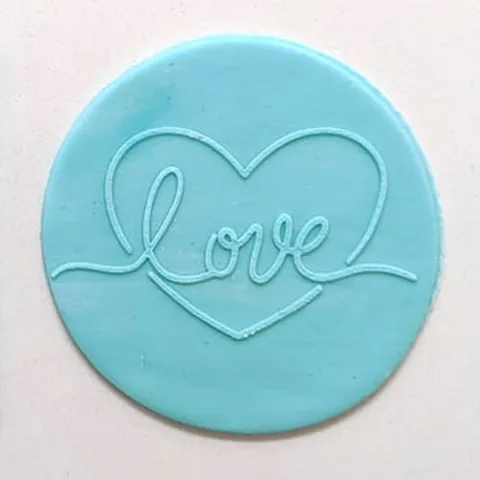A round light blue fondant plaque featuring the word 'love' in a cursive script that forms a heart outline, set against a plain off-white background.