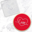 A round red cookie with a bright red center featuring a white line-art heart and the word 'love' in script, shown alongside a clear acrylic debosser tool with a matching design on a white marble surface.