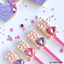 Four heart-shaped decorated lollipop treats with pink and lavender 3D effects and faceted gem accents on pink sticks, arranged on a white surface with scattered pink and white heart sprinkles. A heart-shaped cookie with 'love' is partially visible in the top left.