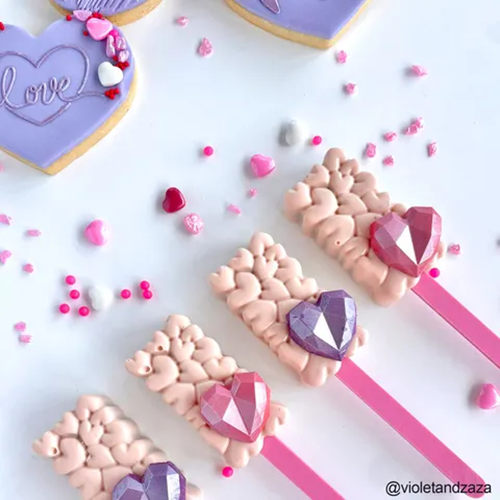 Four heart-shaped decorated lollipop treats with pink and lavender 3D effects and faceted gem accents on pink sticks, arranged on a white surface with scattered pink and white heart sprinkles. A heart-shaped cookie with 'love' is partially visible in the top left.