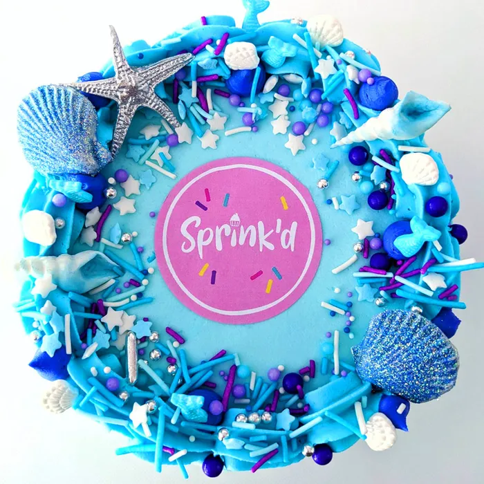 A cake decorated with seashells and mermaid pool party sprinkles in blue silver and white