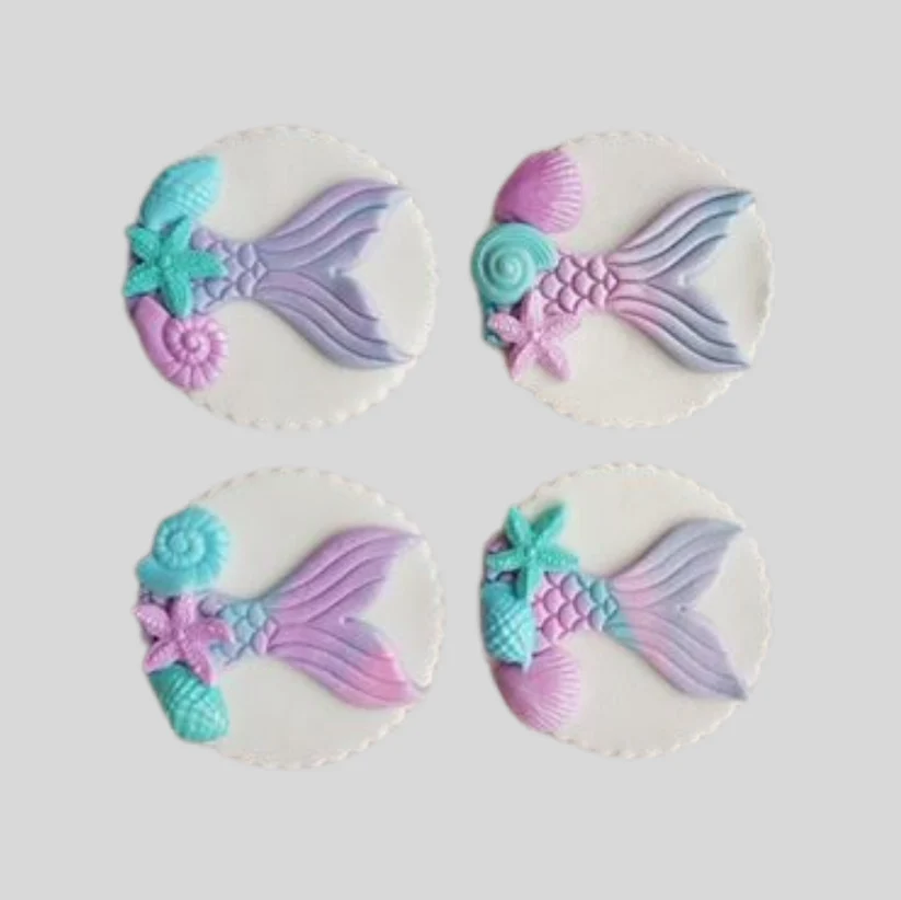 Four mermaid-themed cake toppers or decorations arranged in a 2x2 grid on a light gray background. Each decoration features a pastel mermaid tail in lavender and pink, a teal starfish, a pink starfish, and a shell motif on a white circular backing.