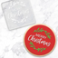 A round Christmas-themed cookiewith a red icing background, white 'Merry Christmas' script, and green leaf wreath decoration, shown alongside a clear debosser of the same design on a light marble surface.