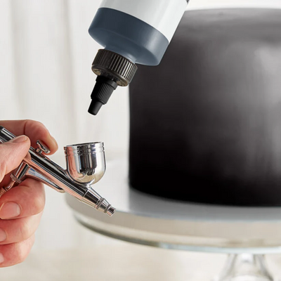 A close-up of a hand holding an airbrush tool while a bottle with a dark applicator tip dispenses material onto a black cake or surface, with a blurred white background.