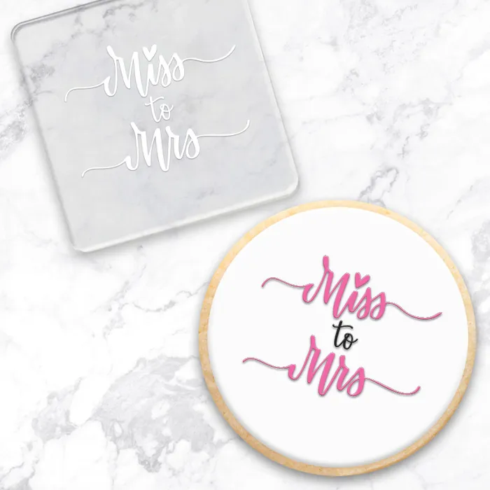 Clear acrylic debosser with 'Miss to Mrs' text . Alo0ngside a finished cookie with the same message, showcasing the tools use - on a marble surface