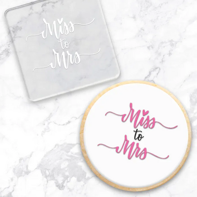 Clear acrylic debosser with 'Miss to Mrs' text . Alo0ngside a finished cookie with the same message, showcasing the tools use - on a marble surface