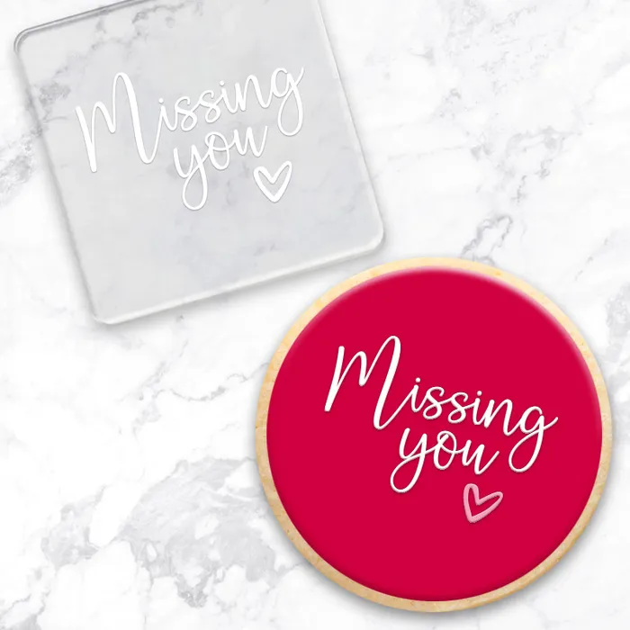 A clear acrylic debosser with the phrase 'Missing you' iand a small heart, shown on a white marble surface. Shown with a round cookies with a bright pink center and natural edge showcasing the design