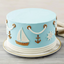A round light blue cake decorated with white rope trim, gold and white anchors, a white sailboat, and a white ship's wheel, shown on a white plate against a neutral background.
