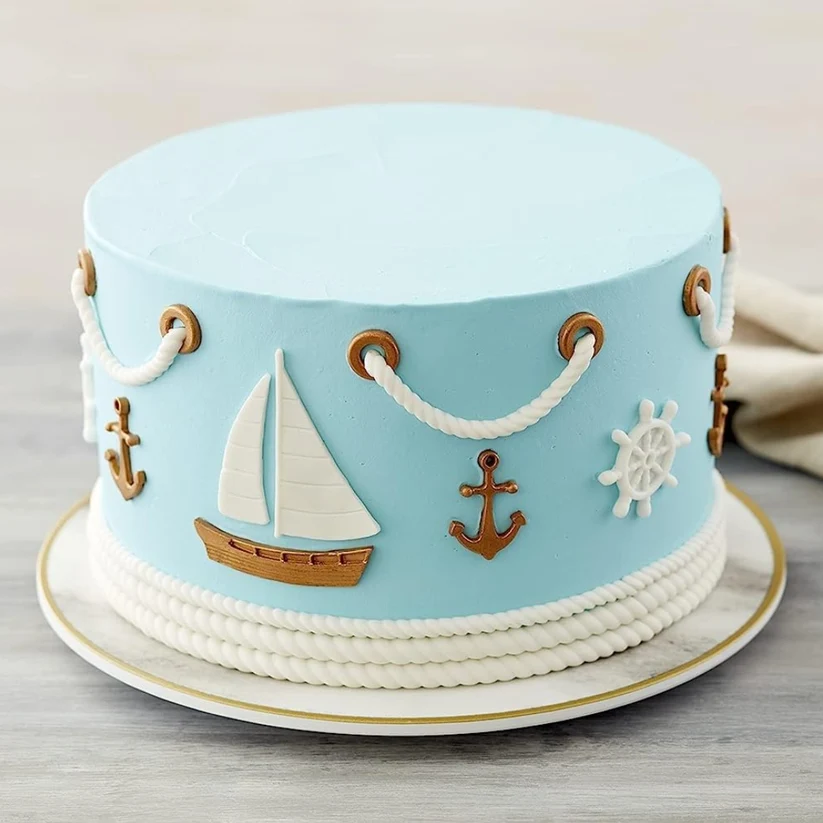 A round light blue cake decorated with white rope trim, gold and white anchors, a white sailboat, and a white ship's wheel, shown on a white plate against a neutral background.