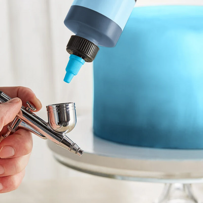 A close-up of a hand holding an airbrush tool while a bottle with a blue nozzle applies liquid to the tip, with a blue frosted cake blurred in the background on a white stand.