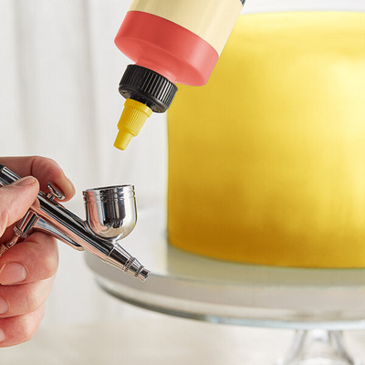 A close-up of a hand holding an airbrush tool while a squeeze bottle with a red and cream body is positioned above it, with a yellow frosted cake on a stand in the blurred background.