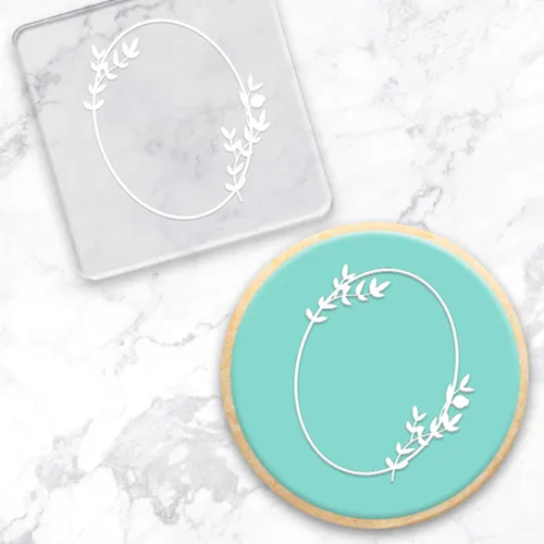 A round frosted cookie with a mint green center and a gold-rimmed edge, decorated with a white oval wreath design, shown next to a matching clear acrylic debosser on a white marble background.
