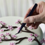A close-up of a hand holding a pair of dark blue curved tweezers, carefully placing a pink cherry blossom flower on a light green iced cake decorated with brown branches.