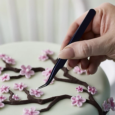 A close-up of a hand holding a pair of dark blue curved tweezers, carefully placing a pink cherry blossom flower on a light green iced cake decorated with brown branches.