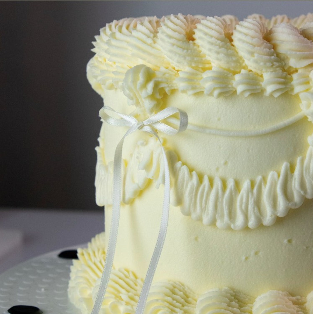 Close-up of a light yellow cake with decorative frosting and a white ribbon against a dark background.