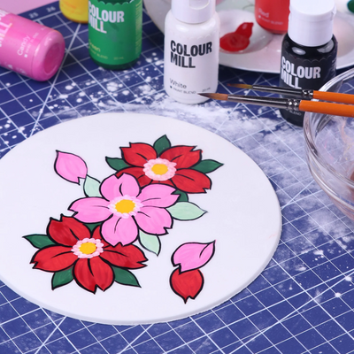Floral painting on round white icing with Colour Mill paint bottles in the background.